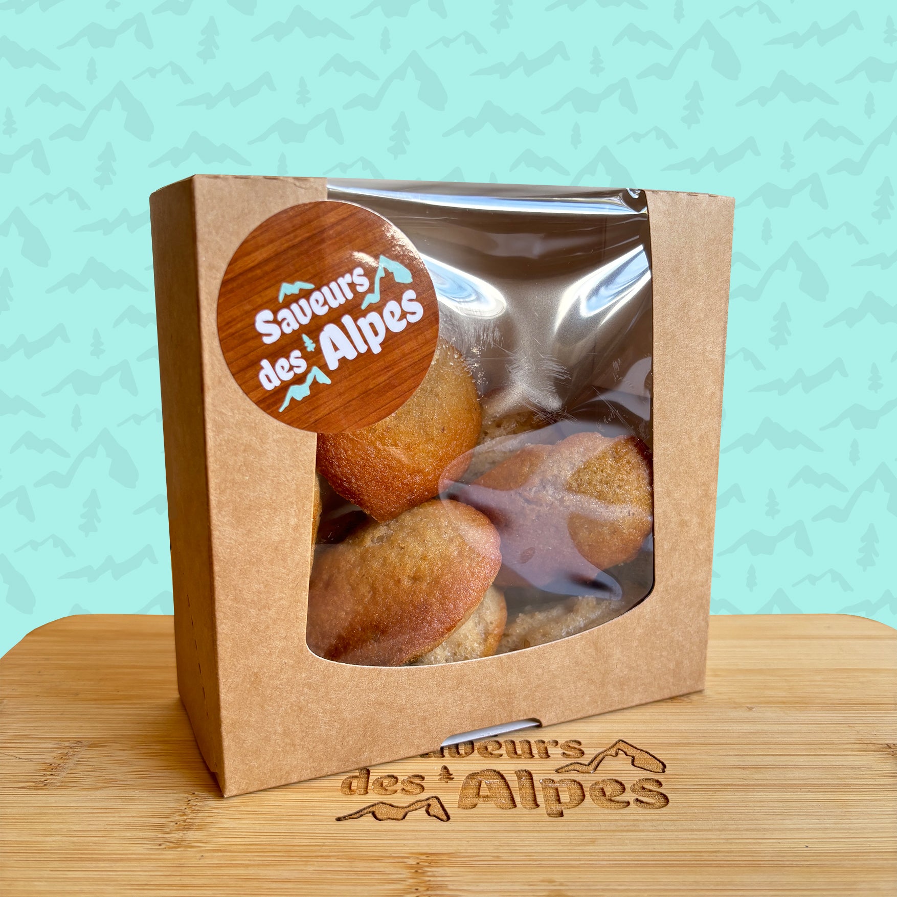 Boîte de madeleines artisanales de La Tourtonnerie – emballage kraft avec étiquette “Saveurs des Alpes”, présentée sur planche en bois gravée, fond bleu à motif de montagnes.