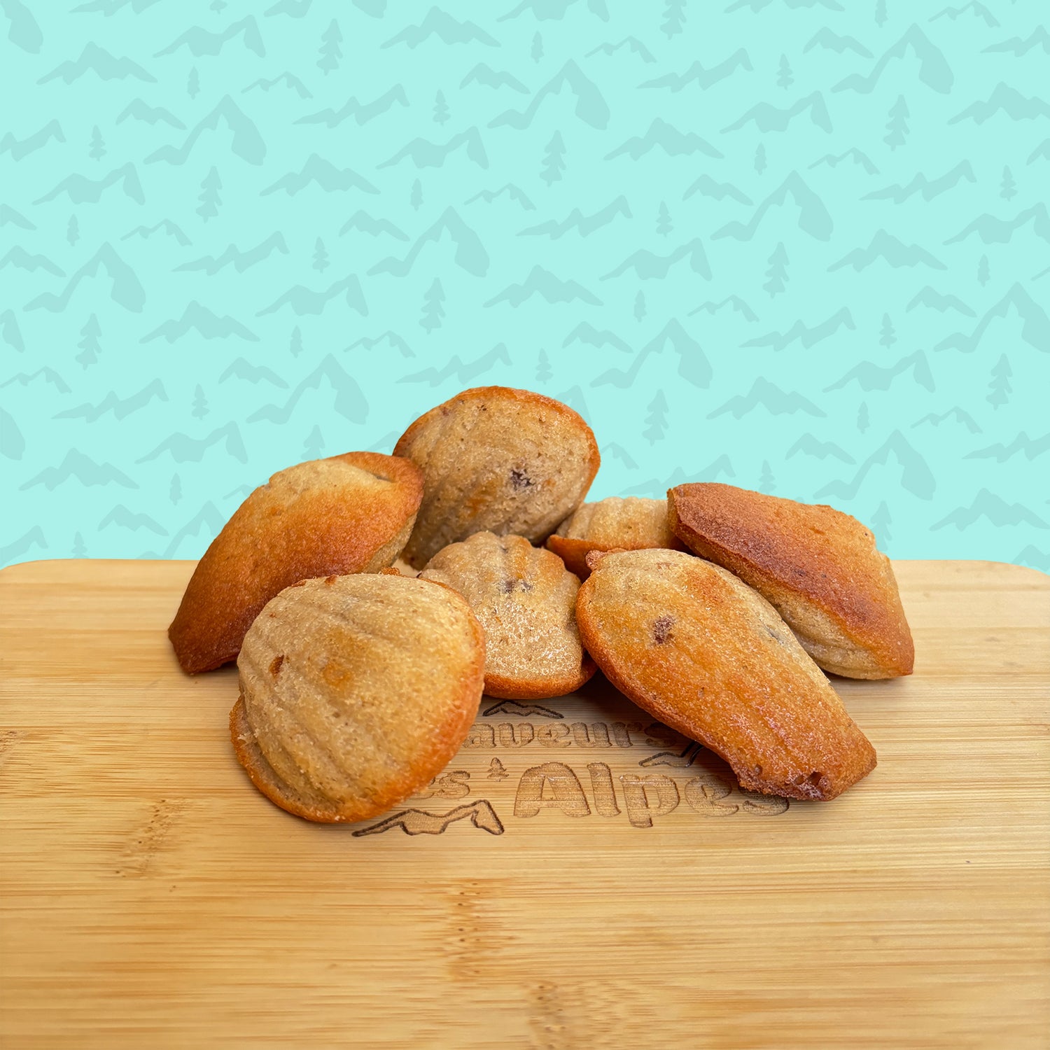 Madeleines artisanales dorées de La Tourtonnerie, disposées sur une planche en bois gravée avec fond bleu illustré de montagnes.